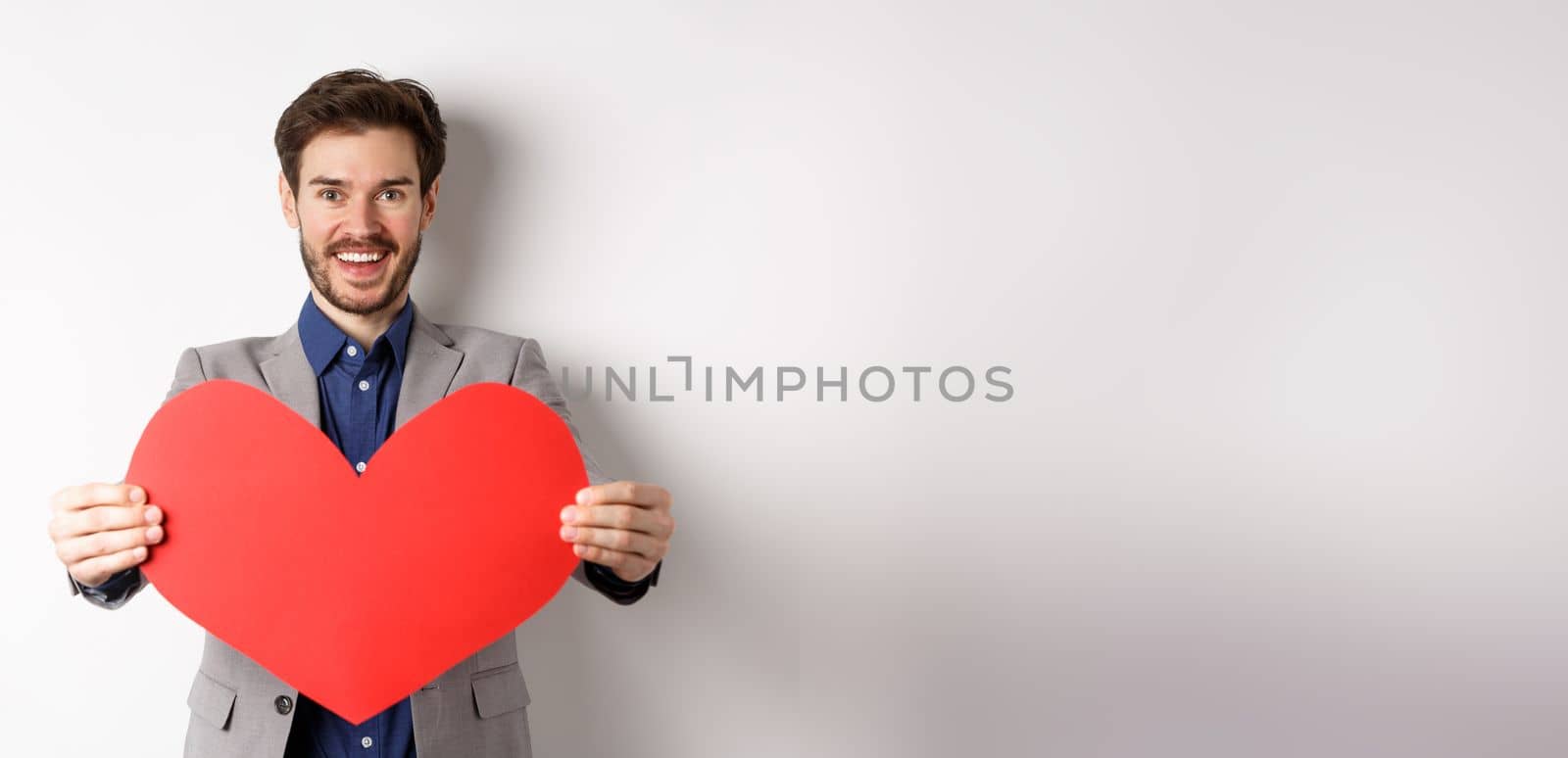 Handsome young man wishing happy Valentines day, giving big red heart sign and smiling, make surprise to lover, standing in suit over white background by Benzoix