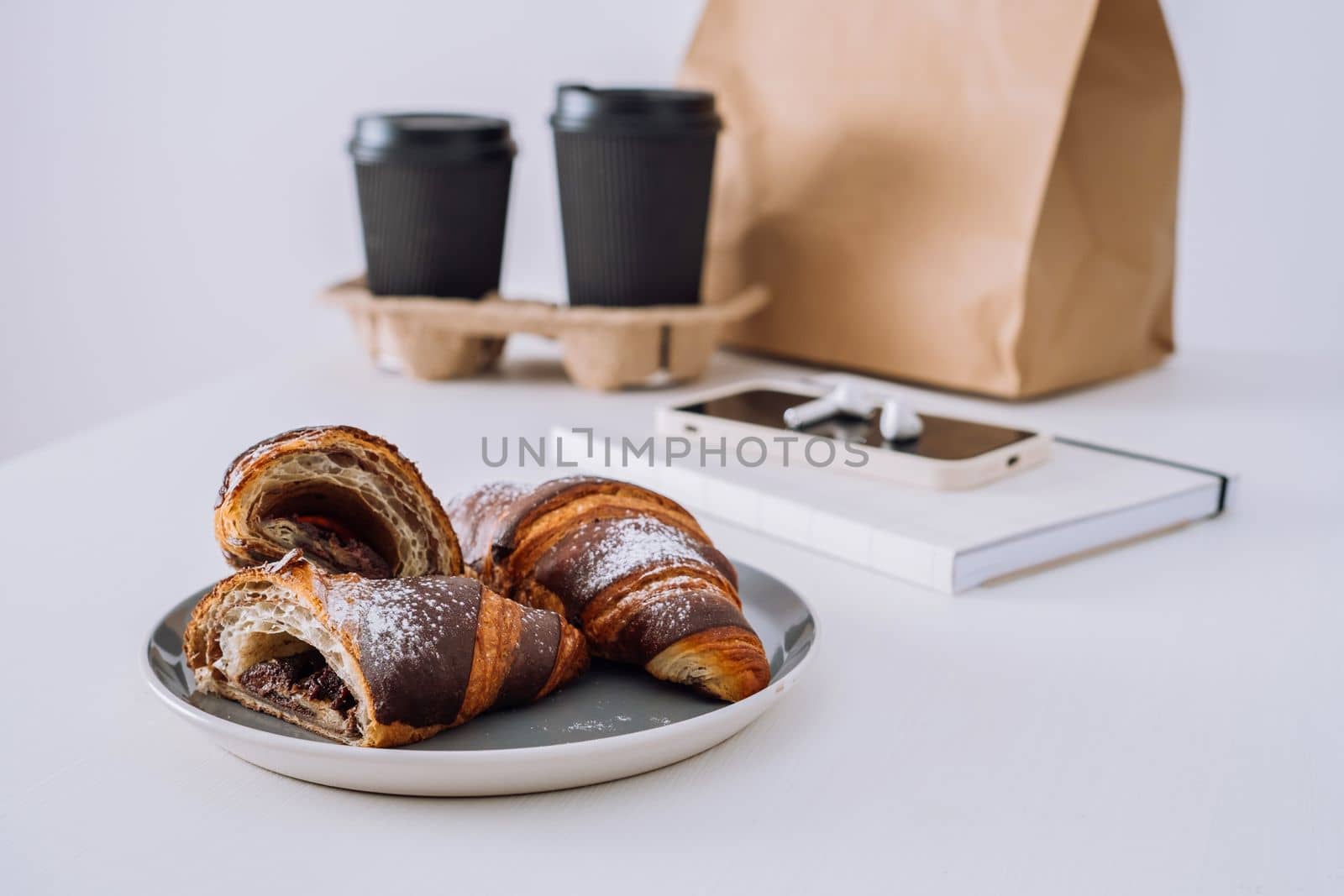 Chocolate croissants with cups of coffee and notepad with smartphone and earphones on the white table, food delivery concept by Romvy