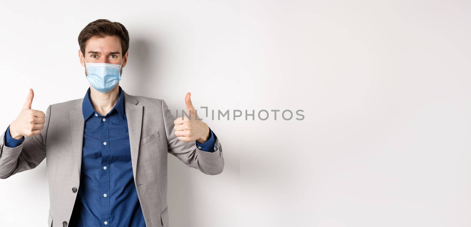 Covid-19, pandemic and business concept. Cheerful man in suit and medical mask using preventive measures in office, showing thumbs up, white background by Benzoix