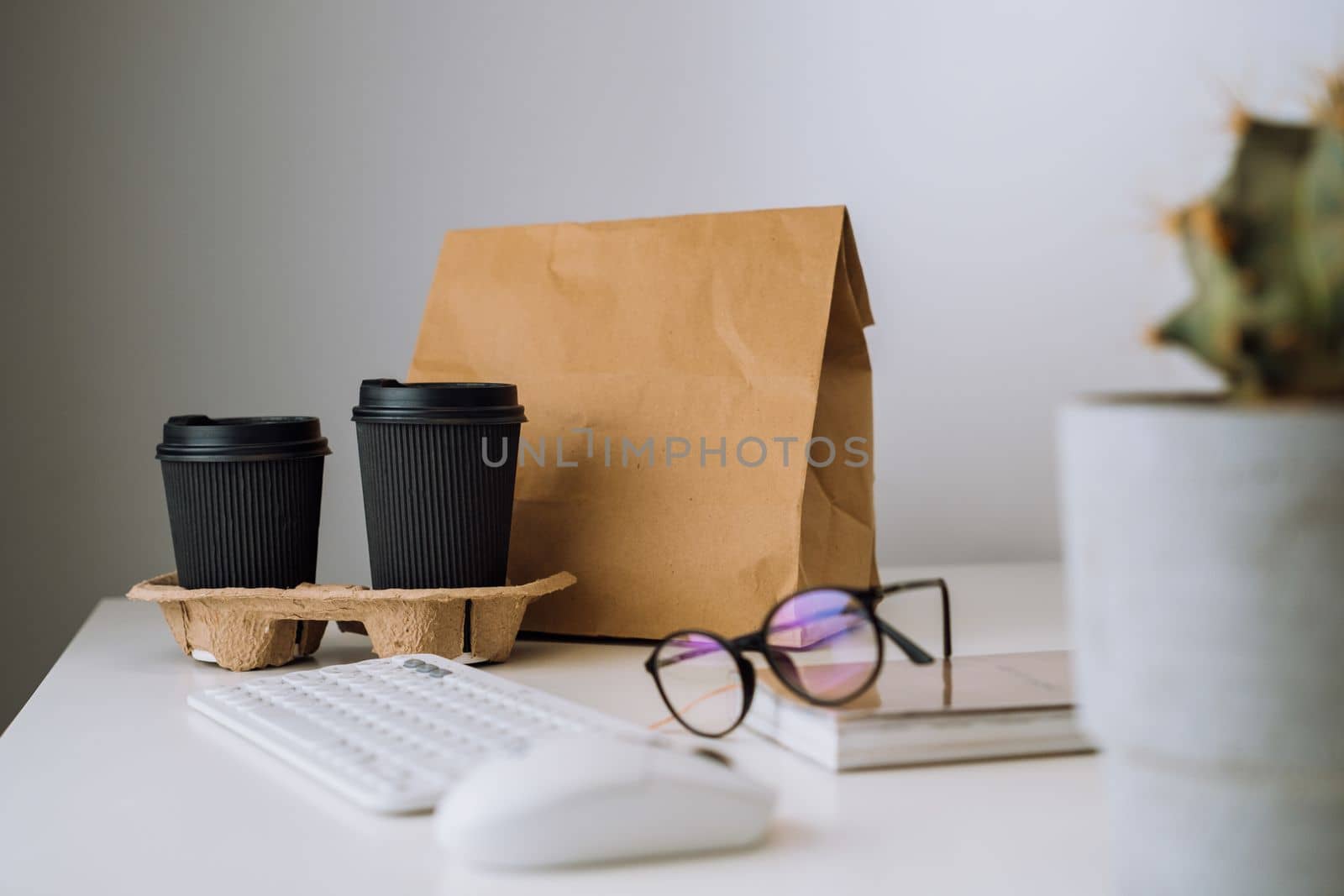 Package with food delivery and cups of coffee at workplace, glasses and notepad with keyboard and cactus on table