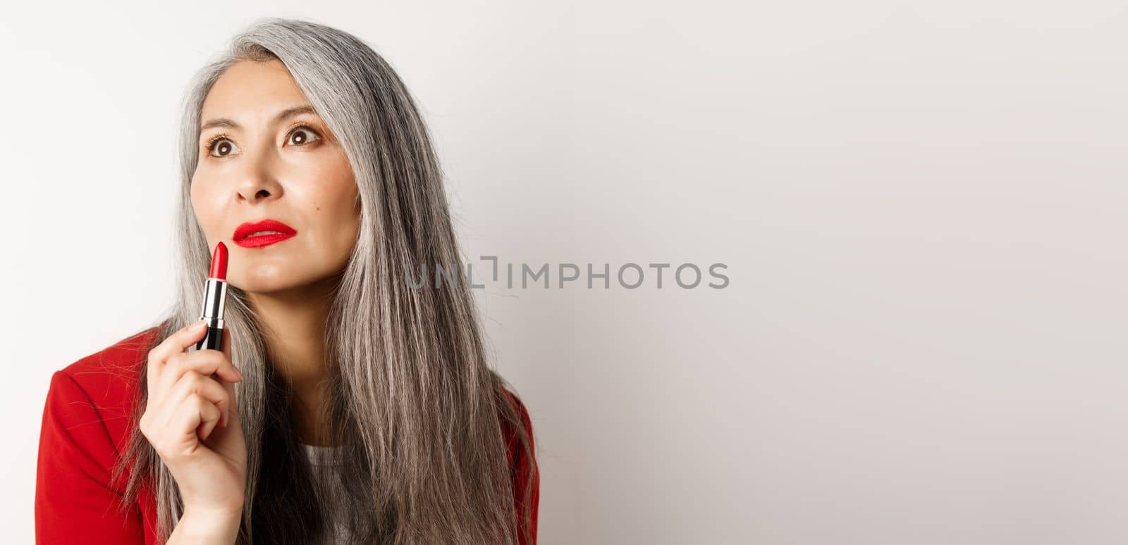 Beauty and makeup concept. Sensual mature asian female with grey hair, looking aside and showing red lipstick, standing over white background by Benzoix