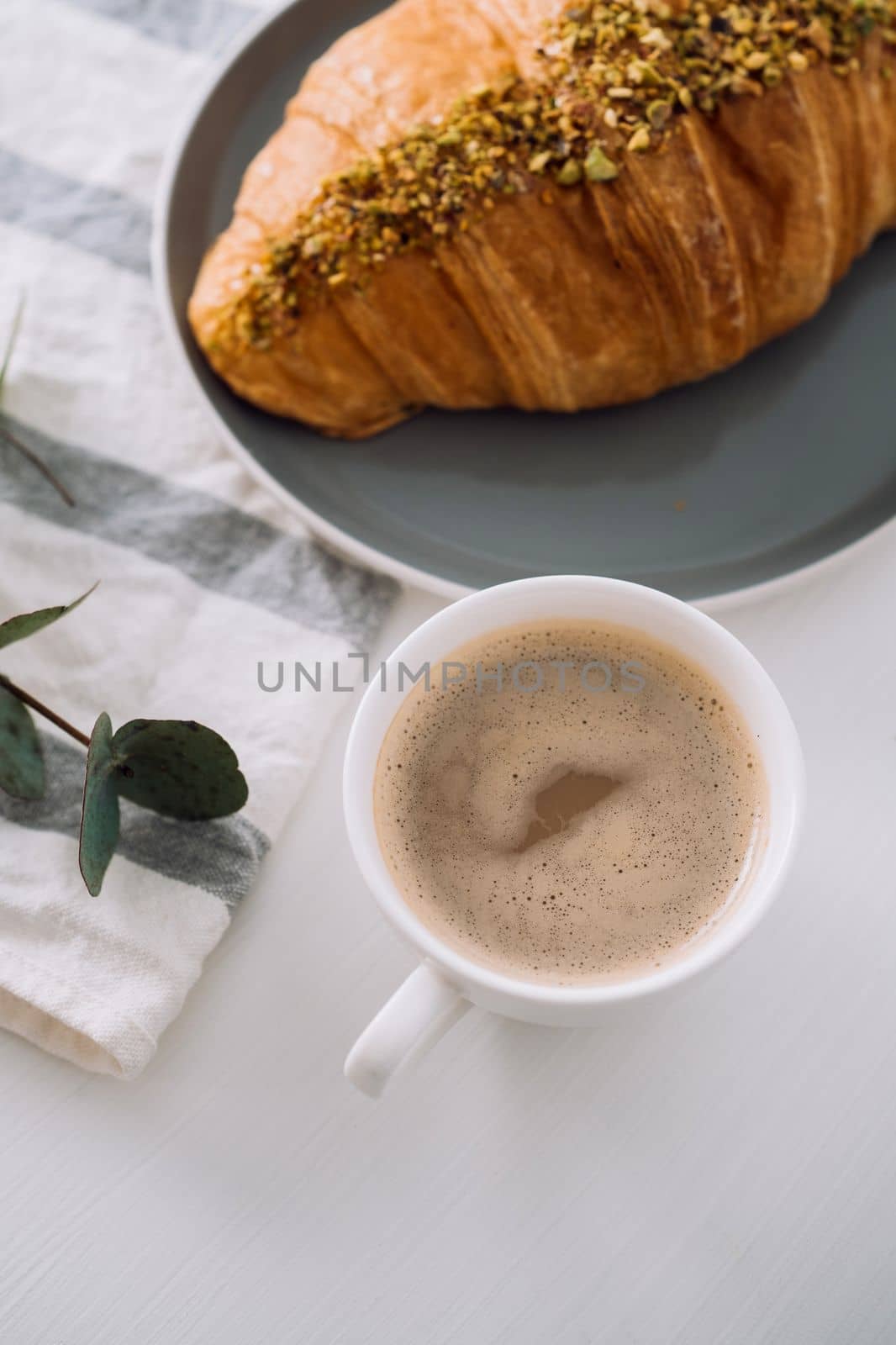 Close up of cup of cappuccino with kitchen towel, croissant and eucalyptus branch on table