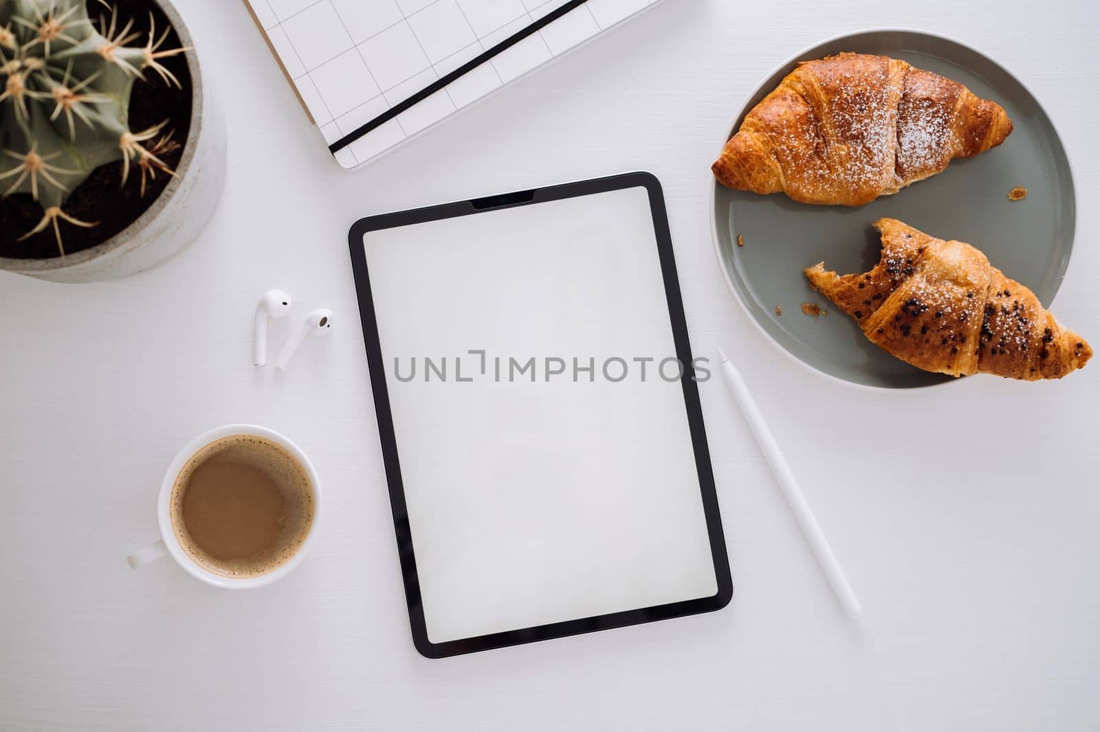 Flat lay of the workplace of the designer illustrator, digital tablet with white screen and stylus pen, cactus, coffee with croissants, notepad and earphones on the table by Romvy