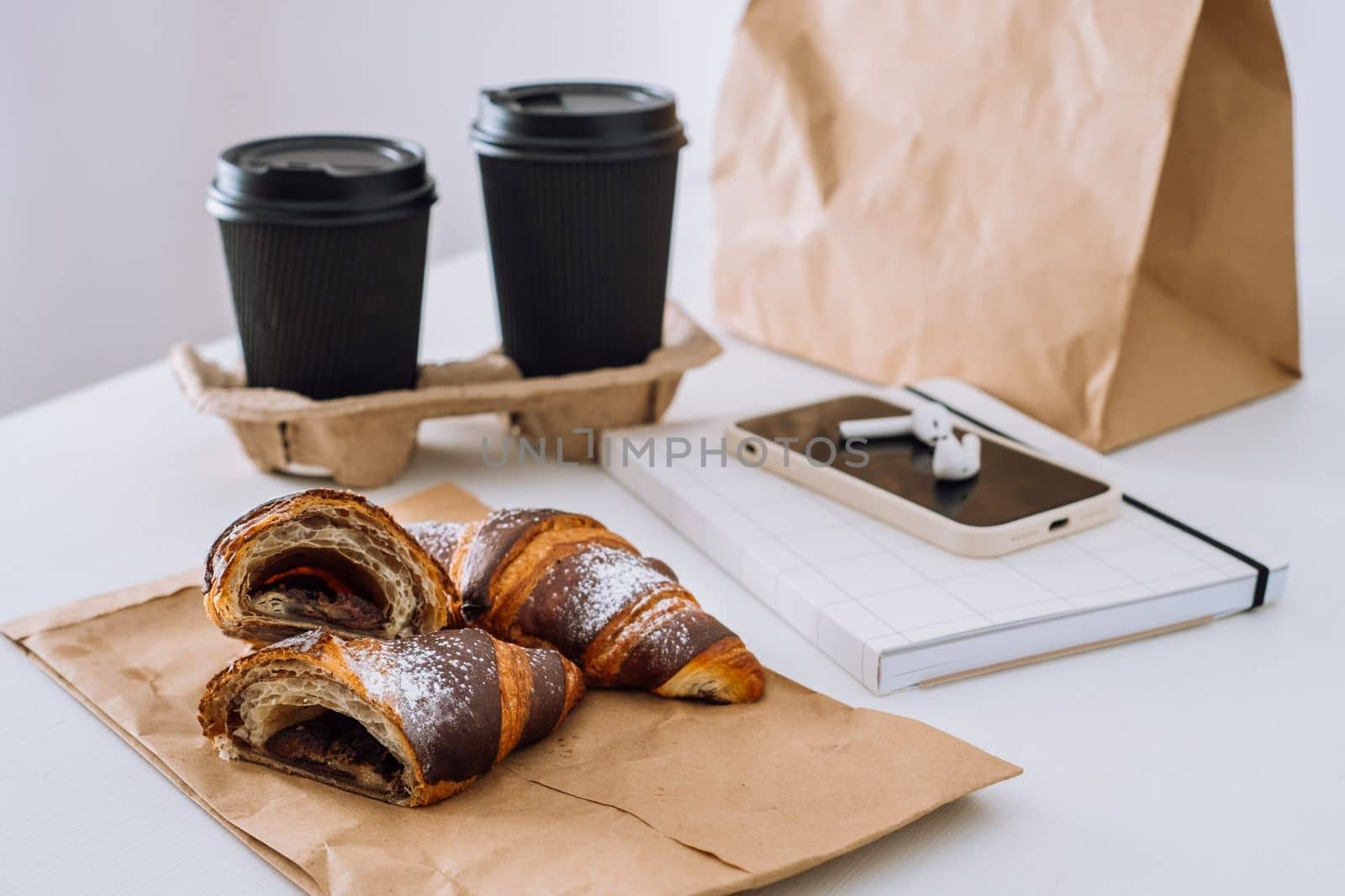 Chocolate croissants with cups of coffee and notepad with smartphone and earphones on white table, food delivery concept