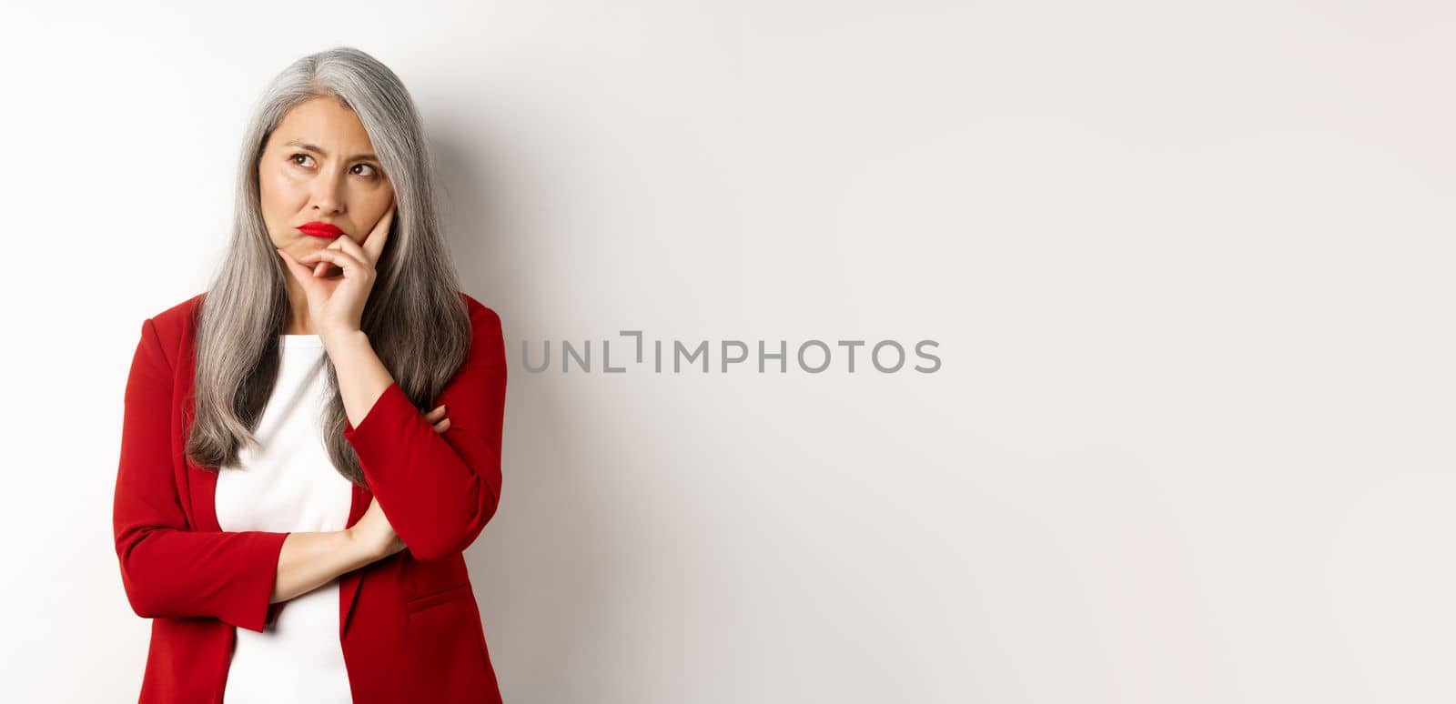 Troubled asian businesswoman in red blazer and lips, pouting and looking left annoyed, standing against white background by Benzoix