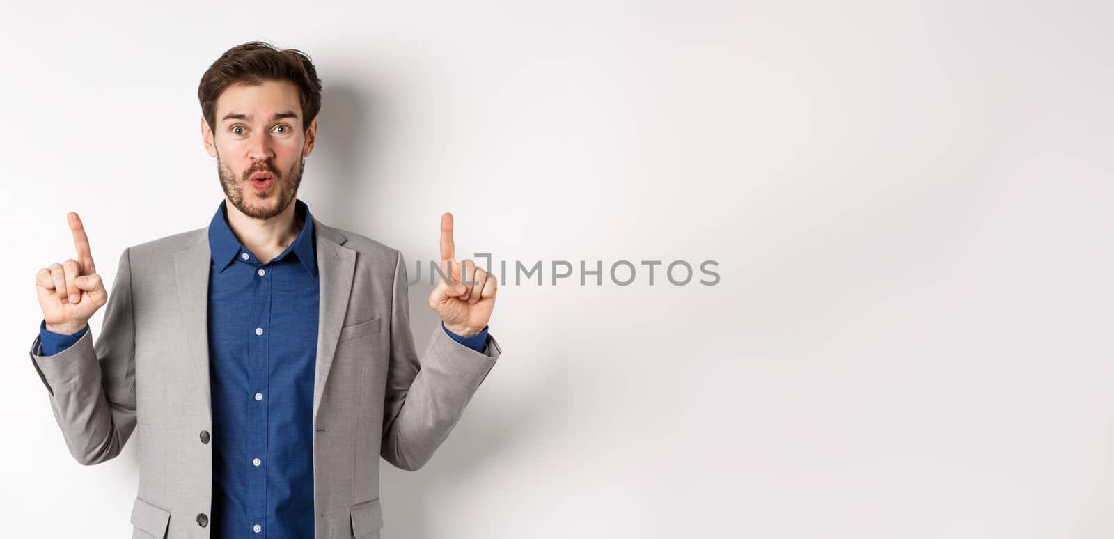 Excited businessman in suit say wow and smiling amused, pointing fingers up at good deal, standing against white background by Benzoix