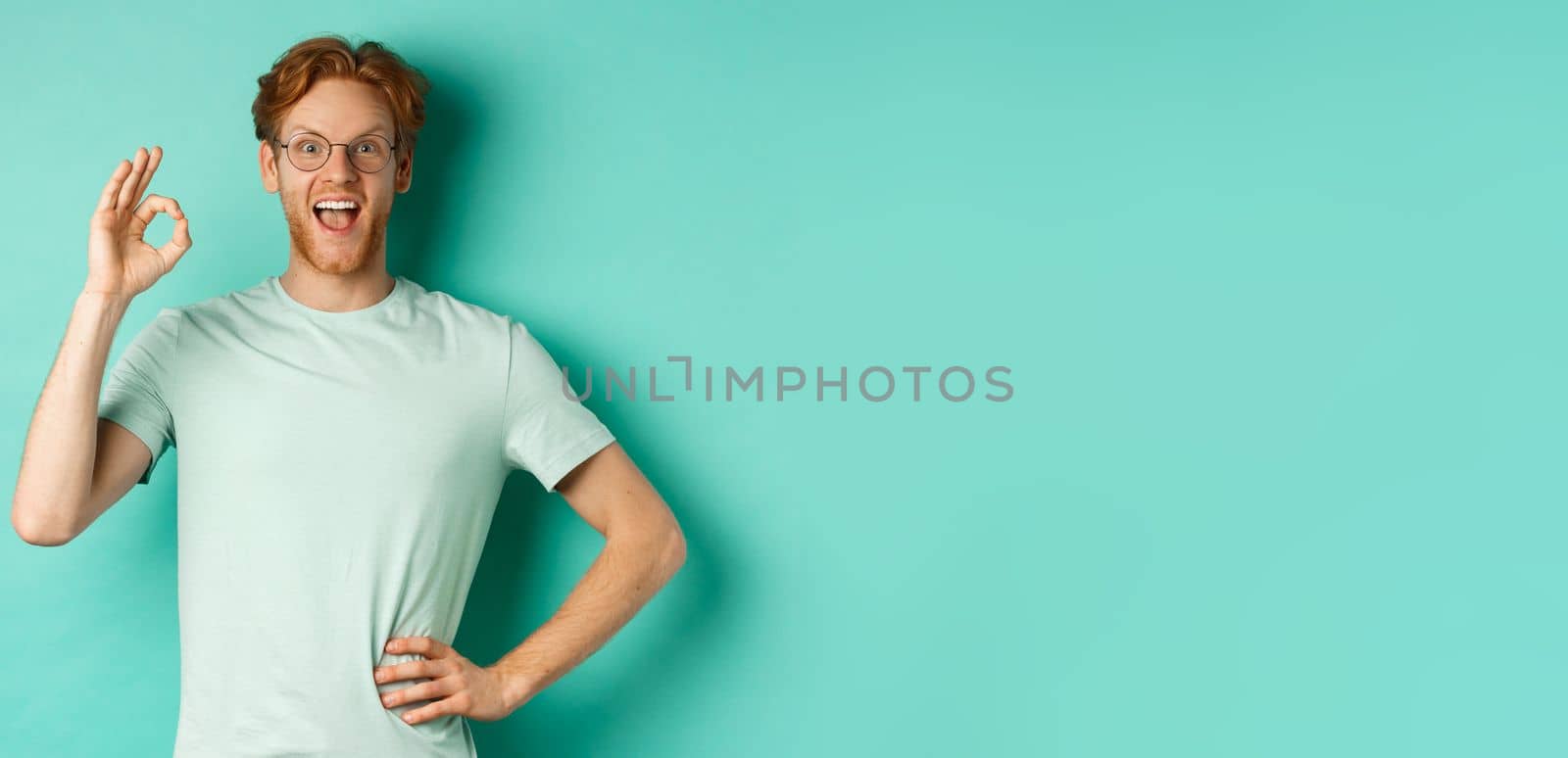 Amused young man with red hair, wearing glasses and t-shirt, showing okay sign and smiling excited, checking out something and approving it, standing over turquoise background by Benzoix