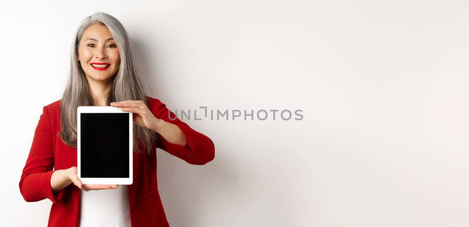 Business. Proud asian female manager showing blank digital tablet screen, smiling and demonstrating promo, standing over white background by Benzoix