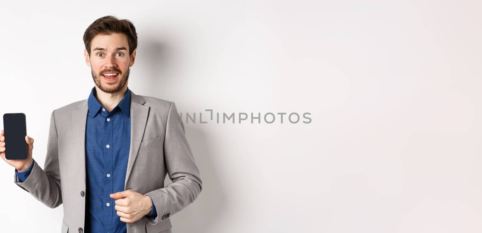 Excited businessman ceo in suit showing empty phone screen and smiling, advertising online promo, standing on white background.