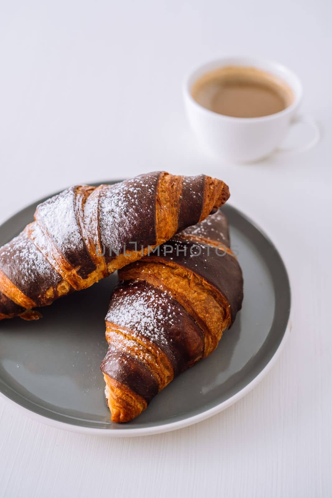 Close up two chocolate croissants on plate with cup of cappuccino in the background