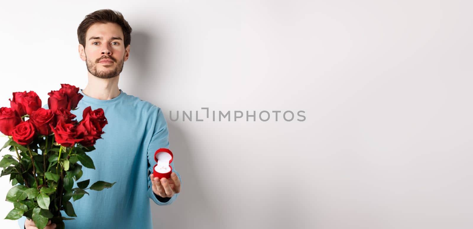 Valentines and relationship. Romantic man boyfriend holding red roses and showing engagement ring, making a proposal on lovers day, standing over white background.