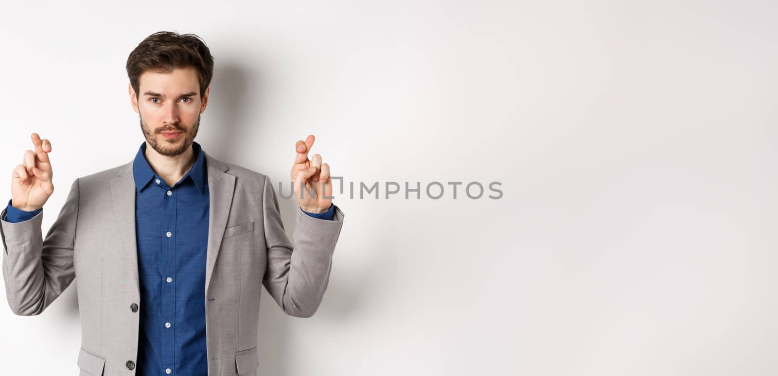 Hopeful man in suit cross fingers for good luck and looking confident in win, feeling determined, standing on white background.