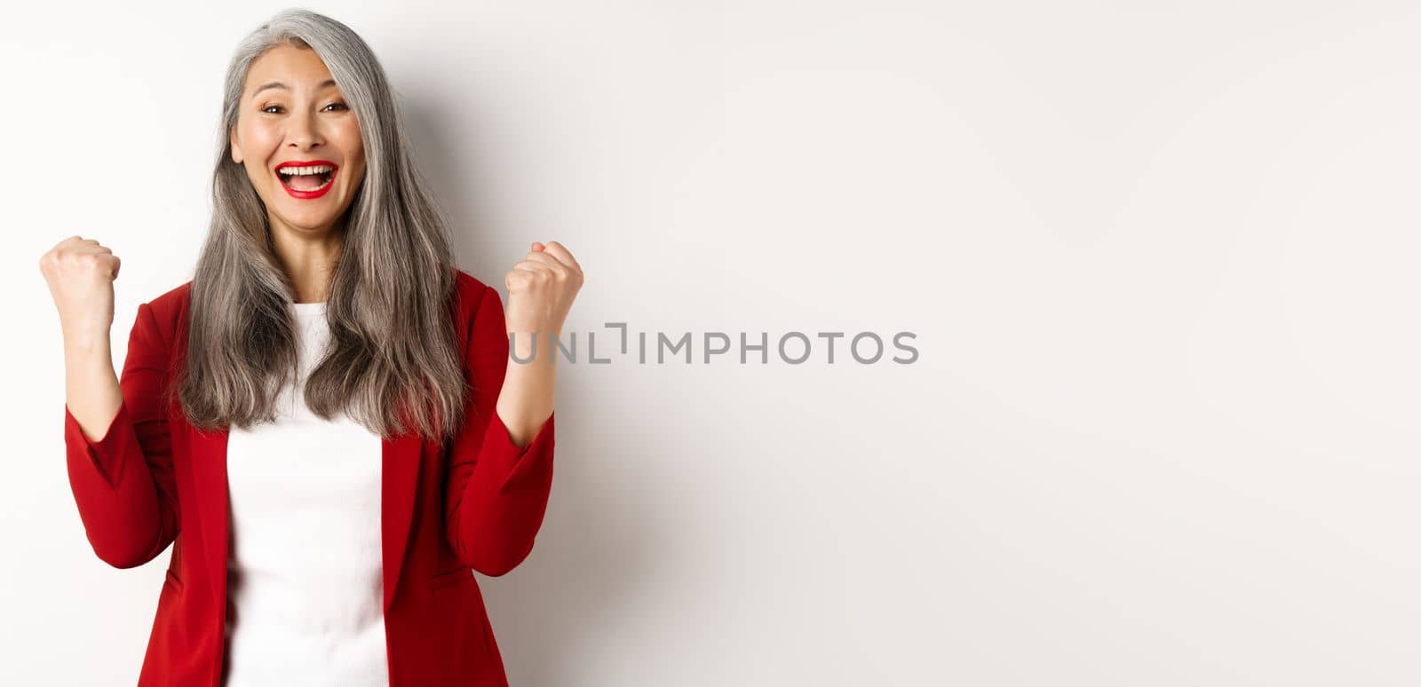 Lucky asian businesswoman achieve success, winning prize and celebrating, saying yes with fist pumps, standing happy against white background.
