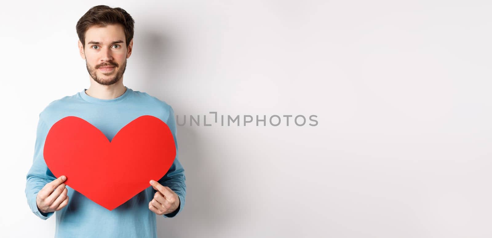 Smiling young man holding valentines heart cutout and looking at camera, waiting for true love girlfriend, standing over white background by Benzoix