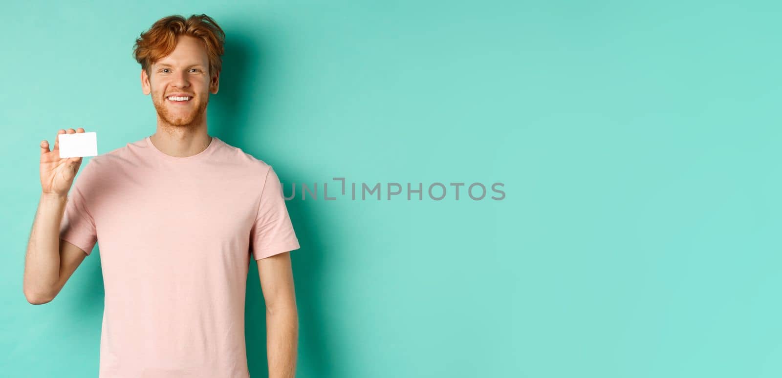 Handsome young man smiling and showing plastic credit card, standing in t-shirt against turquoise background.