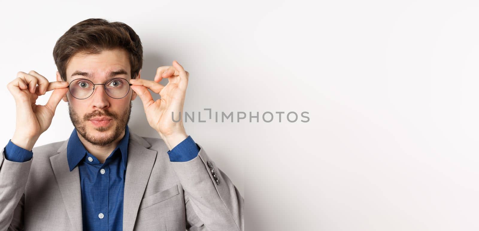 Close-up of handsome man in suit trying put on new glasses, standing on white background.