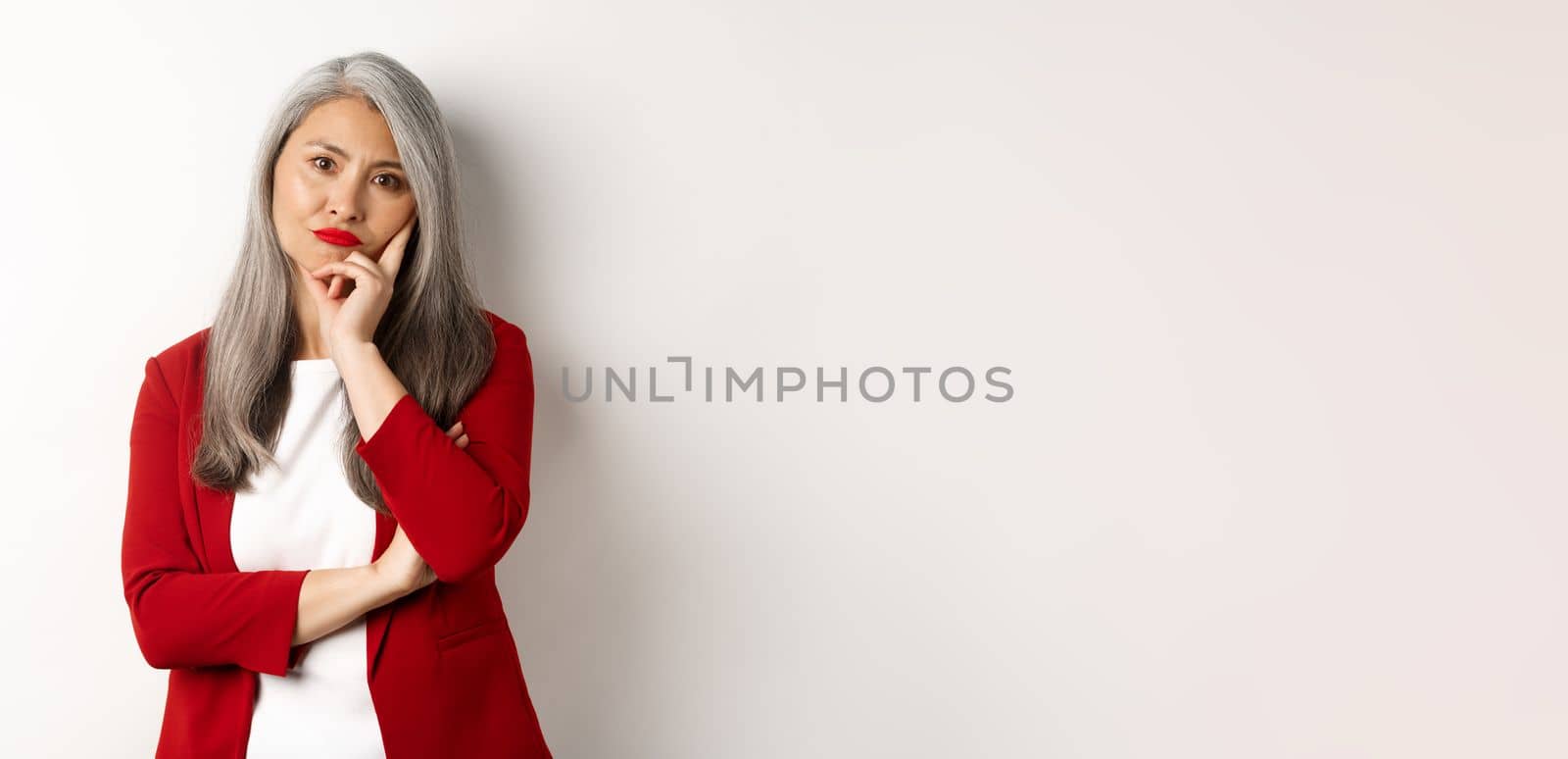 Annoyed and bothered asian businesswoman in red blazer, pouting and looking irritated at camera, standing over white background by Benzoix