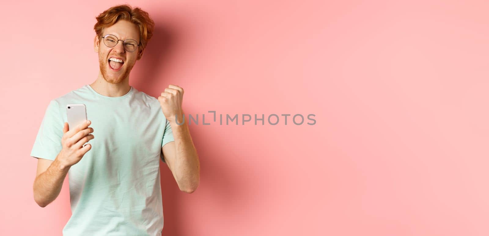 Happy redhead guy in glasses and t-shirt winning online prize, shouting yes with joy and satisfaction, holding smartphone and making fist pump, pink background by Benzoix