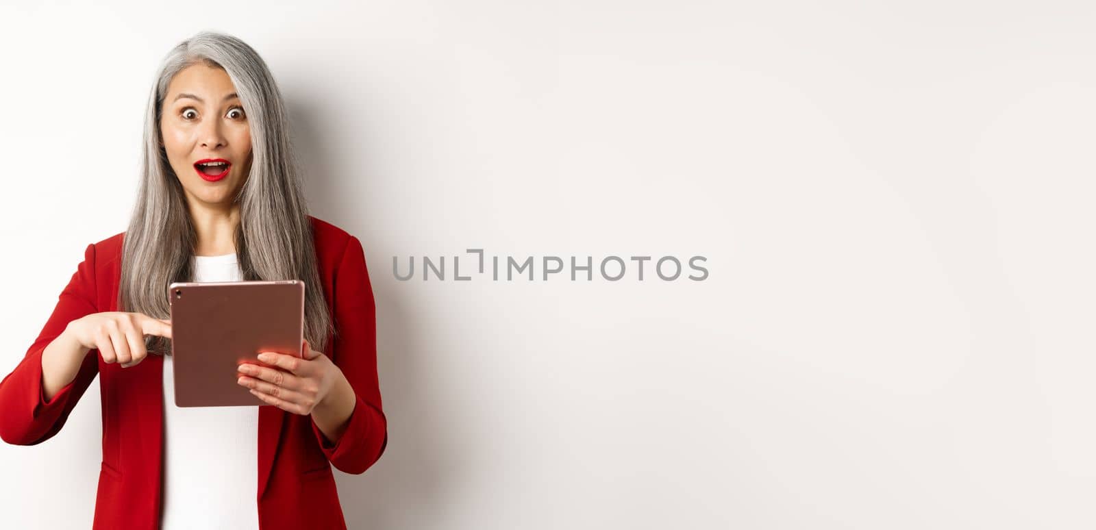 Business. Surprised female senior employer pointing at digital tablet screen, staring amazed at camera, checking out online promotion, white background.