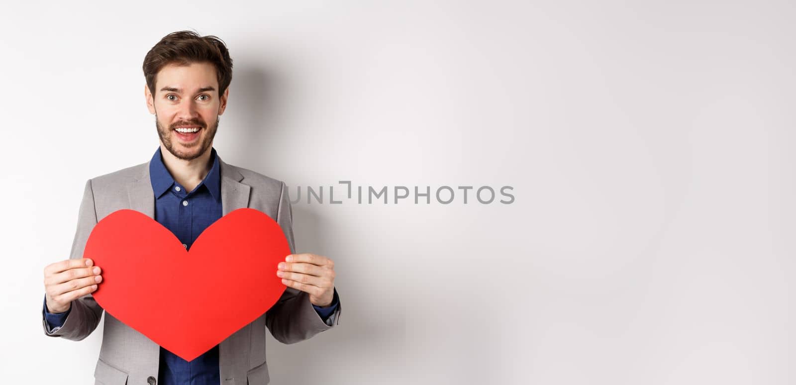 Happy man confessing in love, showing heart cutout and smiling at camera, standing in suit on romantic date with lover, white background.