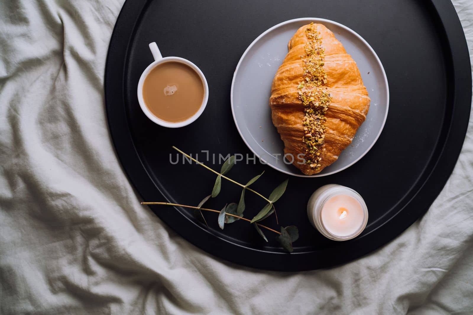 Flat lay of cup of cappuccino, croissant and scented candle with eucalyptus branch on the tray on bed by Romvy