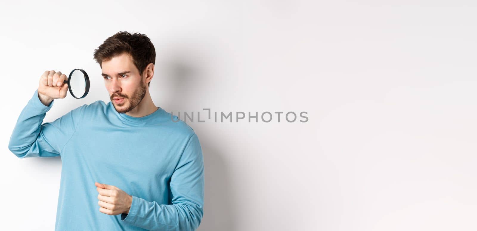 Young handsome man look through magnifying glass with curious face, investigating or searching for something, standing on white background by Benzoix
