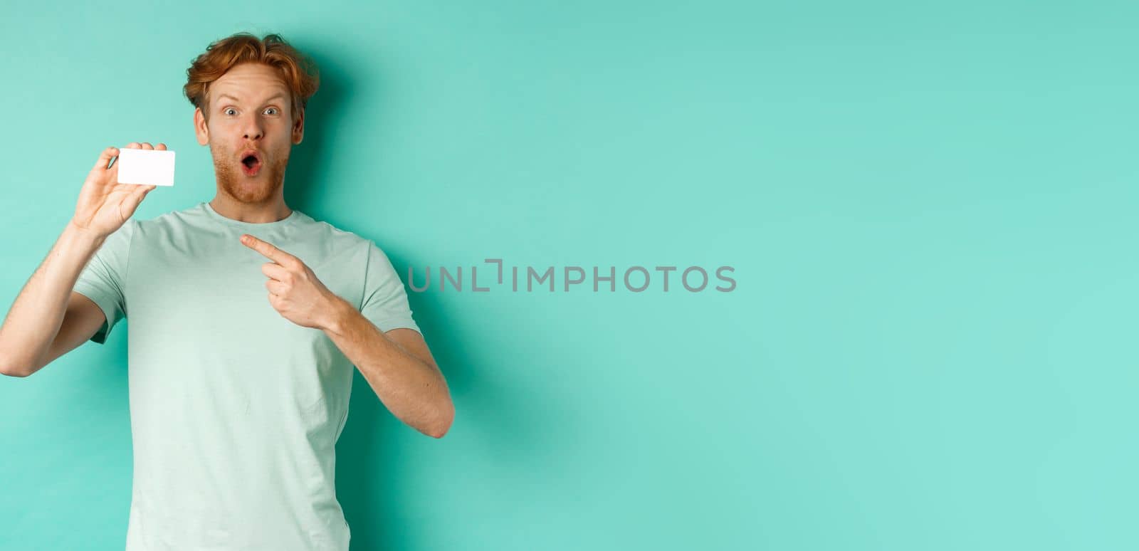 Shopping concept. Handsome redhead man in t-shirt showing plastic credit card and smiling, standing over turquoise background.