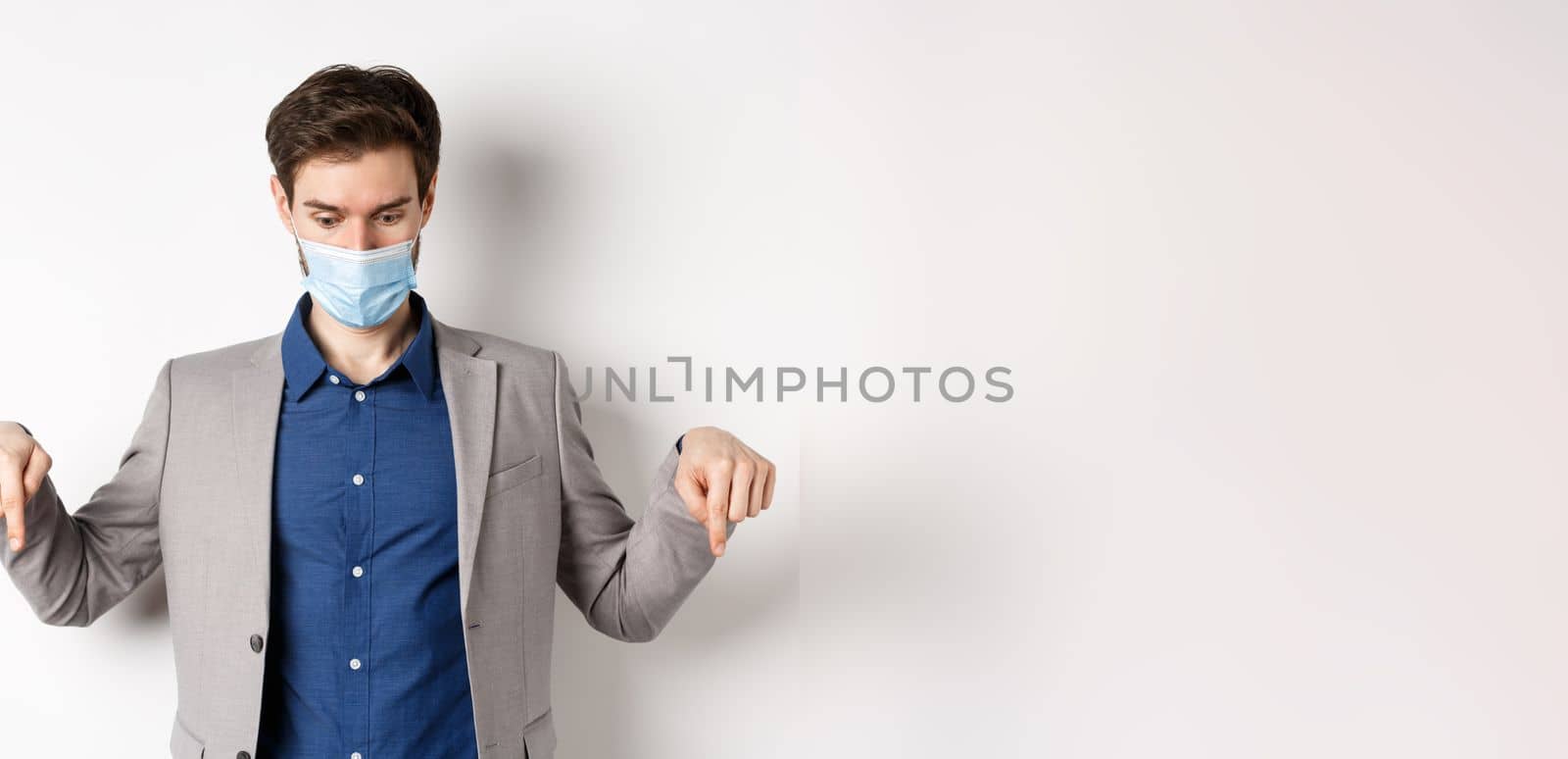 Covid-19, pandemic and business concept. Surprised guy in medical mask and suit looking and pointing down with popped eyes, gasping amazed, white background by Benzoix