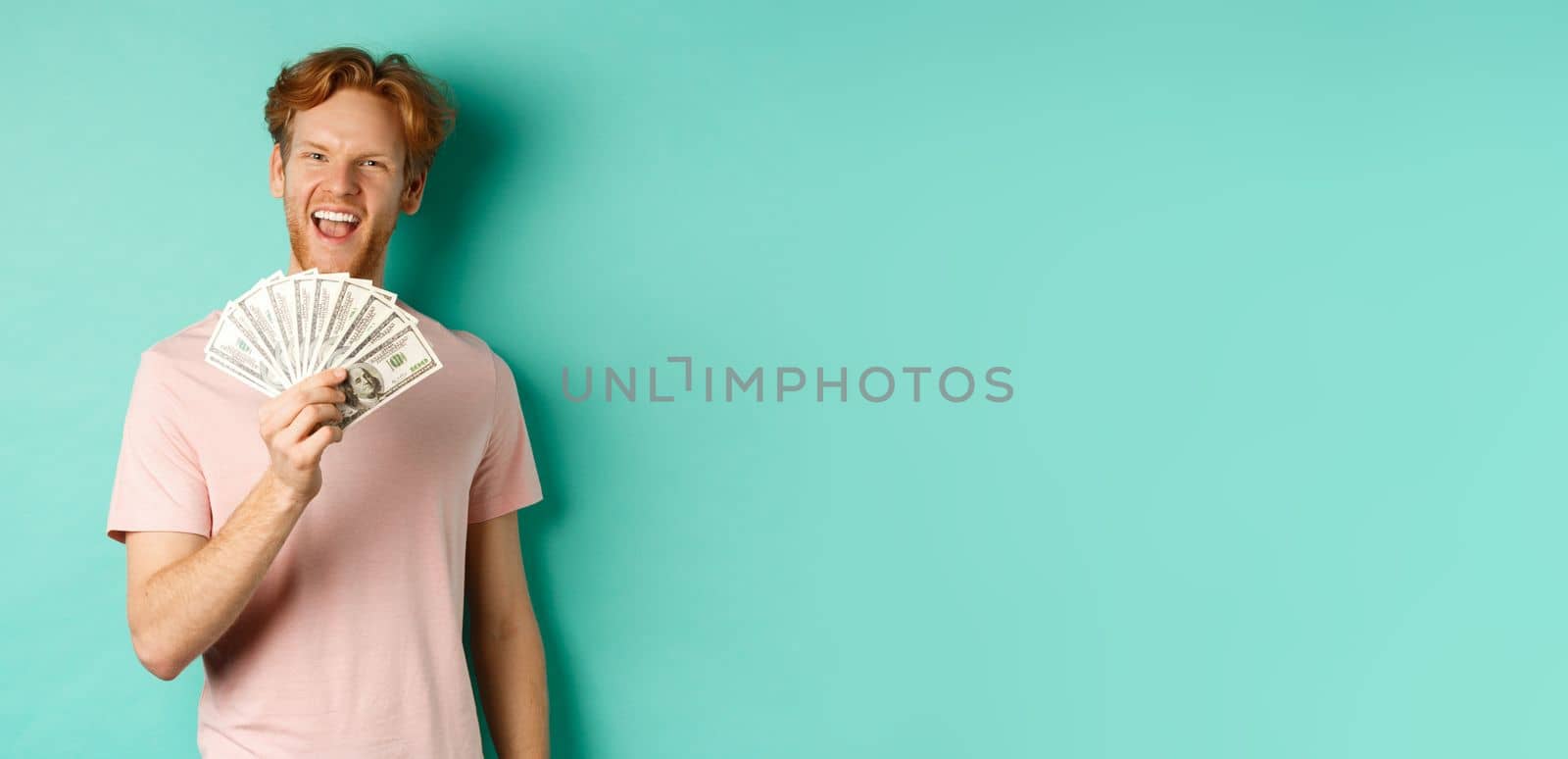 Happy redhead man in t-shirt showing money in dollars and smiling, making smug faces after winning cash, standing over turquoise background by Benzoix