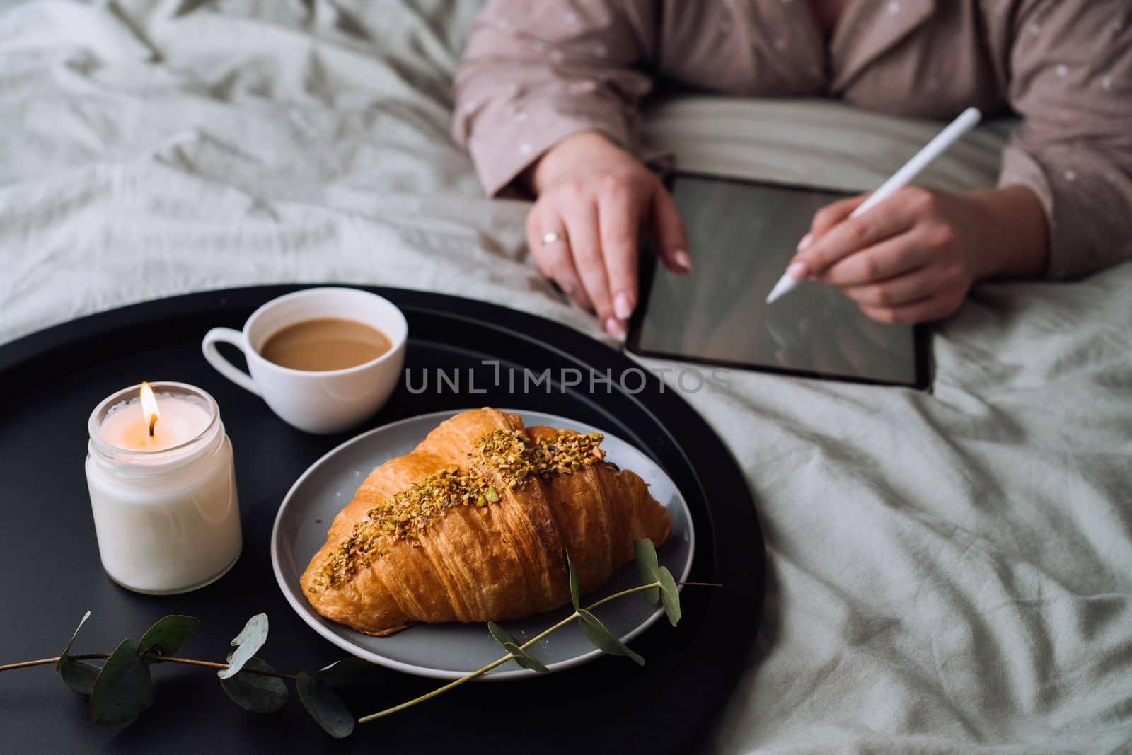 Croissant with cup of cappuccino and scented candle on the tray, woman drawing on digital tablet on background by Romvy