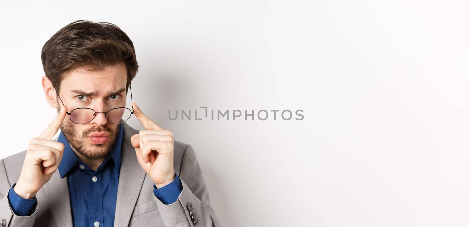 Close-up portrait of suspicious man in suit, taking-off glasses and frowning with hesitant face, standing on white background.