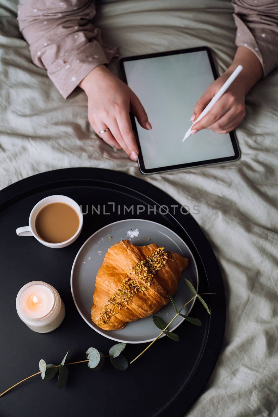 Woman using digital tablet for drawing while lying on the bed and enjoying croissant with cappuccino