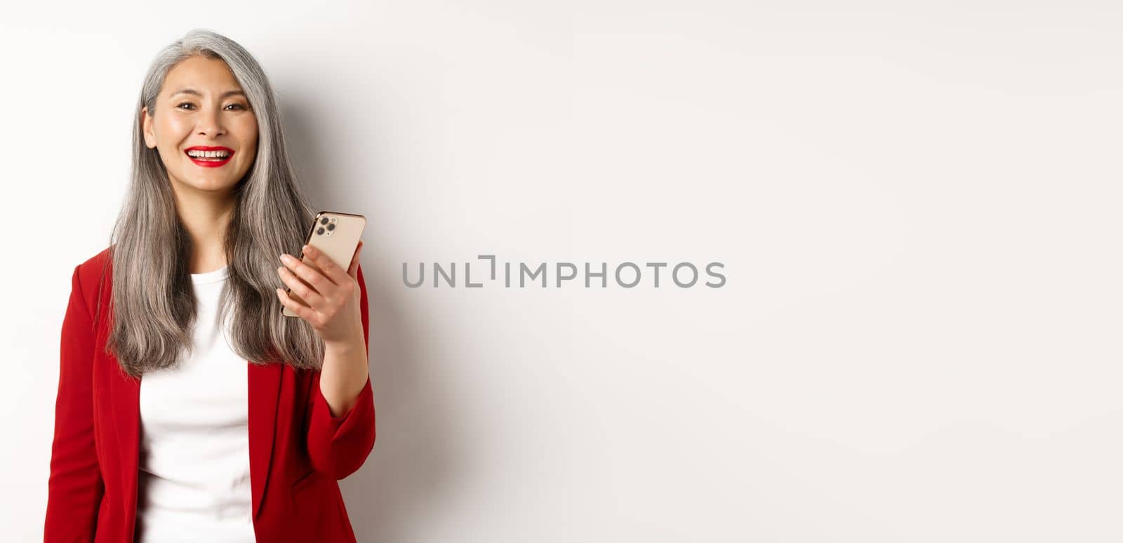 Asian female entrepreneur in red blazer using smartphone, smiling happy at camera, standing over white background.