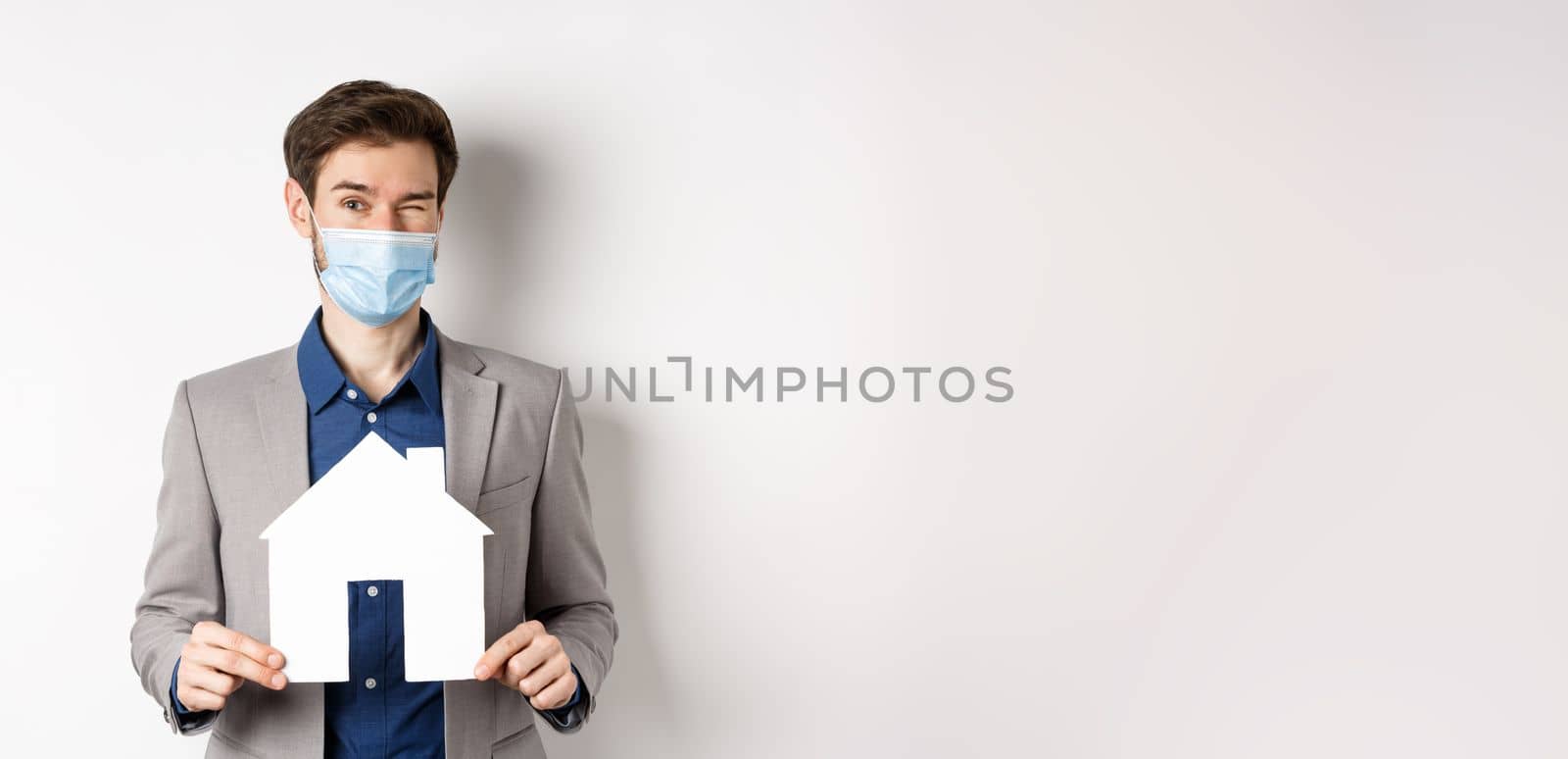 Real estate and covid-19 concept. Cheerful young man in medical mask and suit winking at camera, advertising new flats, showing paper house cutout by Benzoix