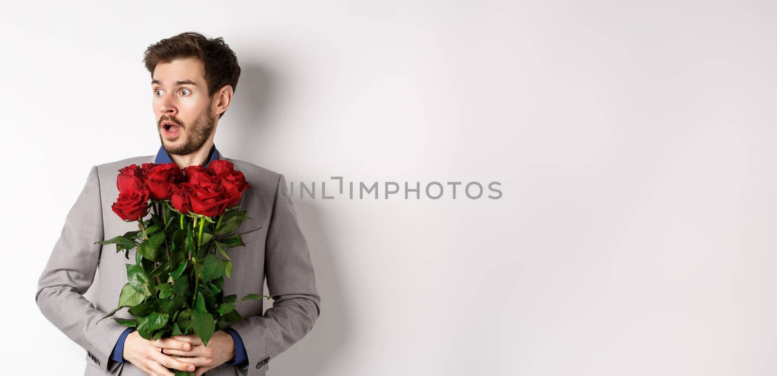 Handsome young man in suit holding red roses, looking left with surprised and startled expression, standing on Valentines day over white background by Benzoix