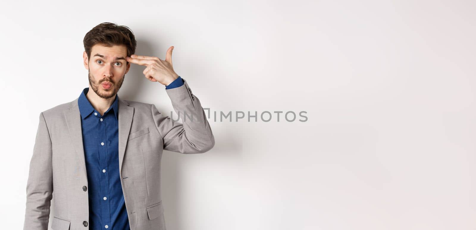 Annoyed guy in business suit shoot himself with hand gun near head, look distressed and tired after work, standing on white background.