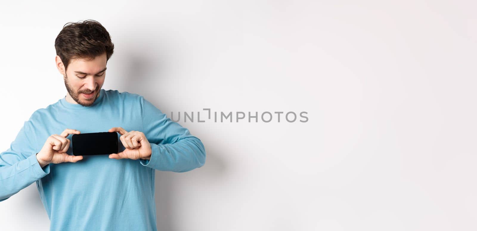 E-commerce and shopping concept. Caucasian man showing blank mobile screen and looking at it, standing curious against white background by Benzoix