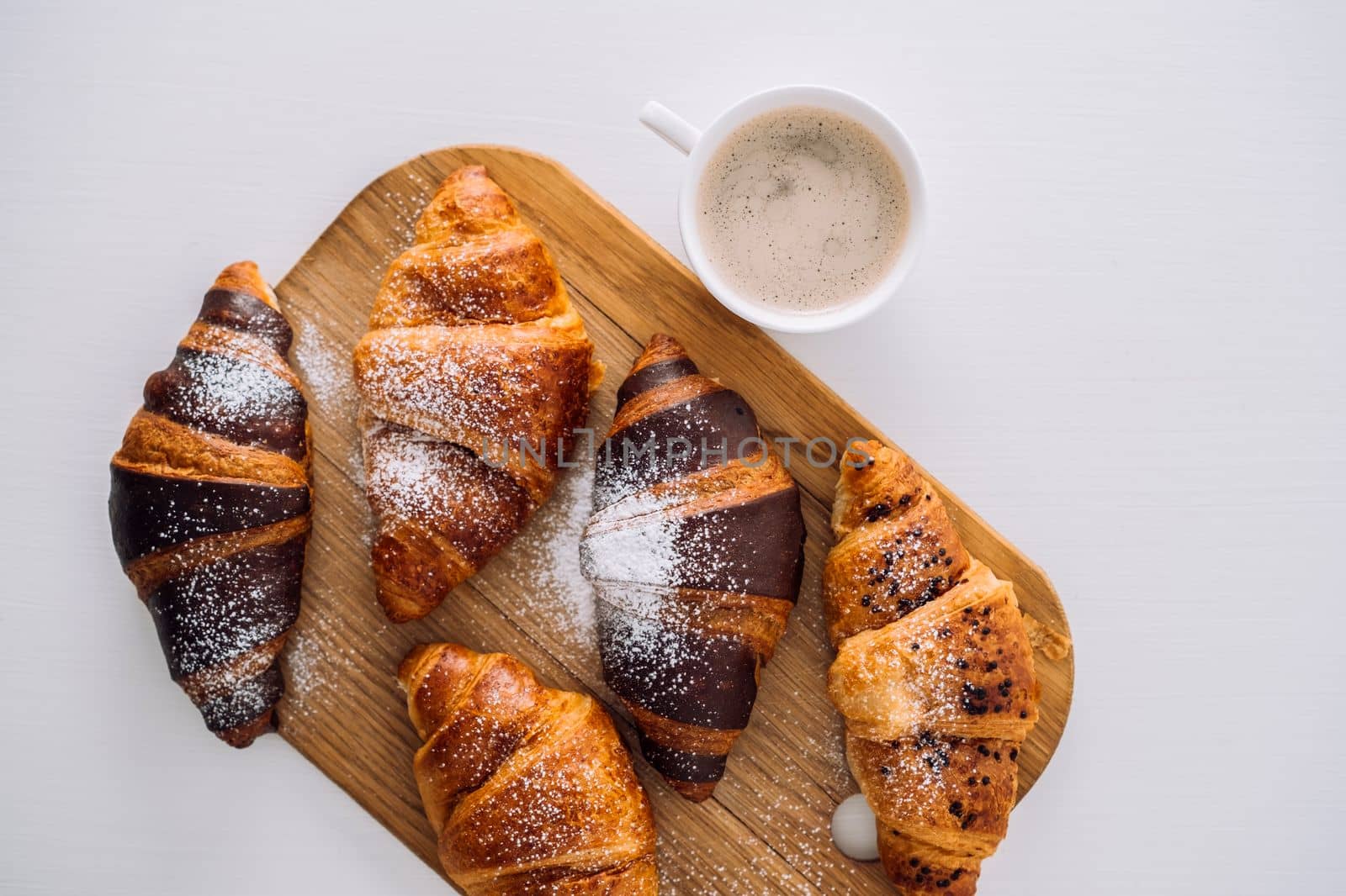 Cup of coffee and bunch of appetizing brown and chocolate croissants with powdered sugar on a wooden board on white table, flat lay