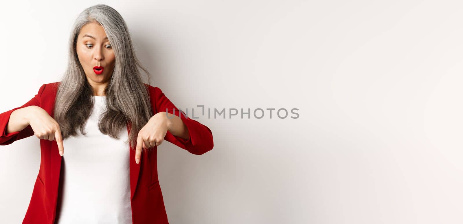 Impressed asian mature woman checking out promo, pointing and looking down with amazed face, standing in blazer over white background by Benzoix