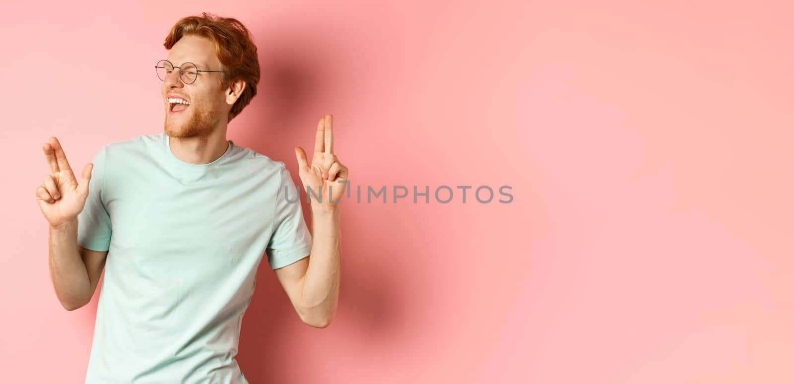 Cheerful redhead guy listening music and dancing, triumphing or celebrating success, standing over pink background by Benzoix