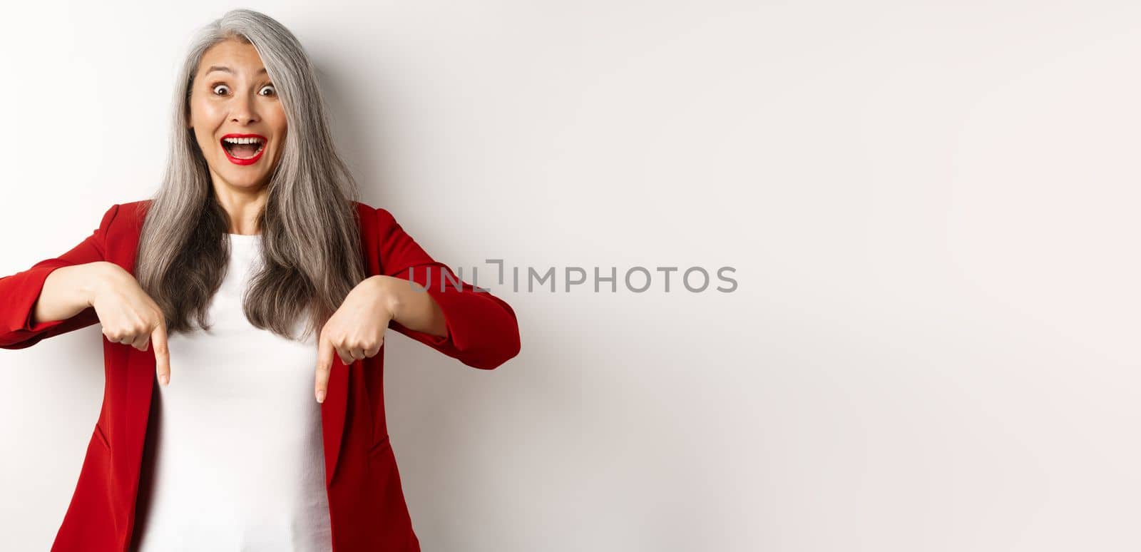 Portrait of happy asian lady in red blazer showing logo, pointing fingers down and smiling cheerful, check this out gesture, white background.