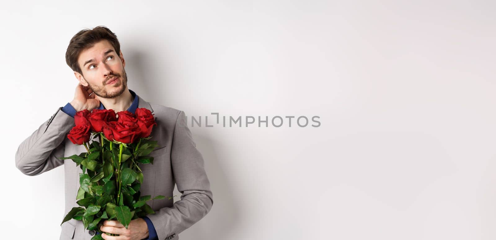 Thoughtful young man in suit going on valentines day date, holding bouquet of roses, thinking and scratching head, standing against white background by Benzoix