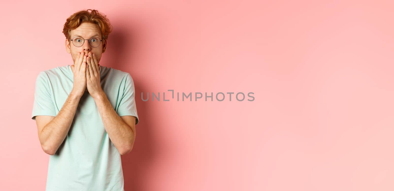 Shocked and speechless redhead guy covering mouth, staring at camera startled, standing in t-shirt against pink background.