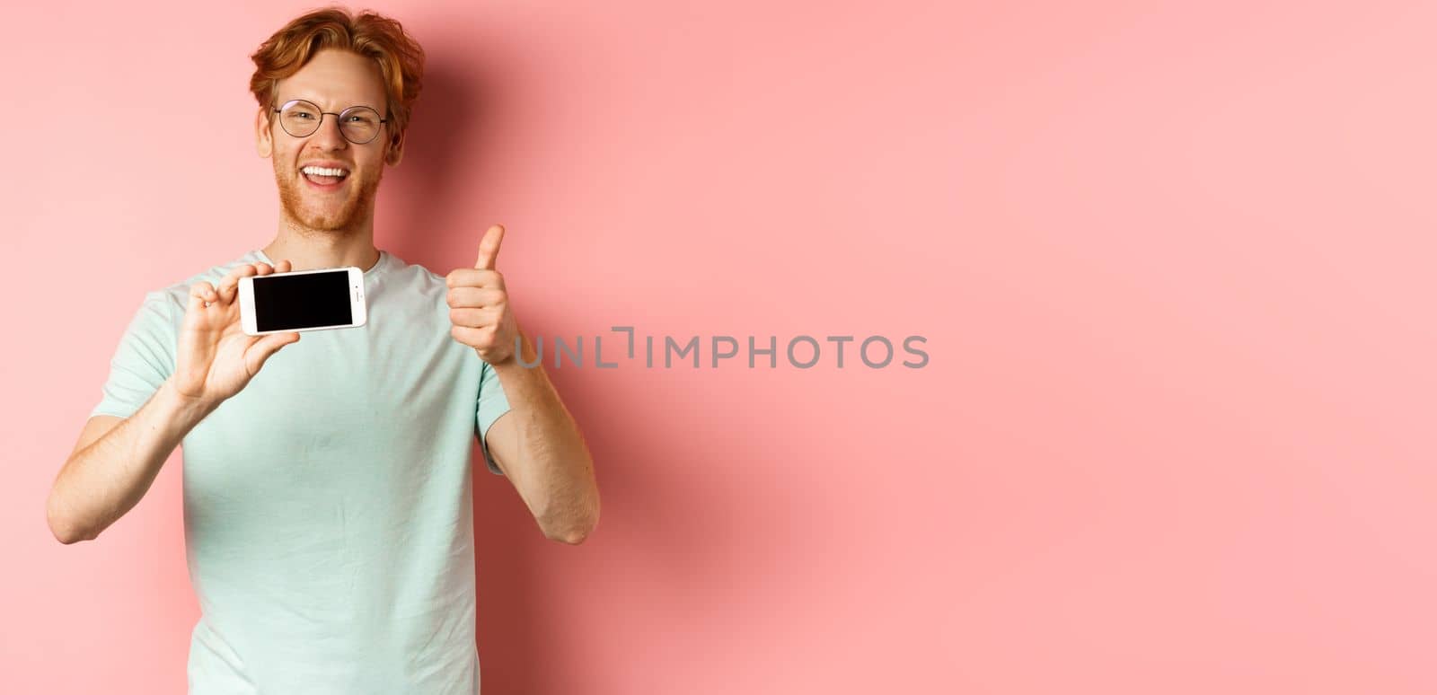 Happy european man with ginger hair and glasses, showing mobile blank screen horizontally and thumbs-up, recommend online promo, pink background.