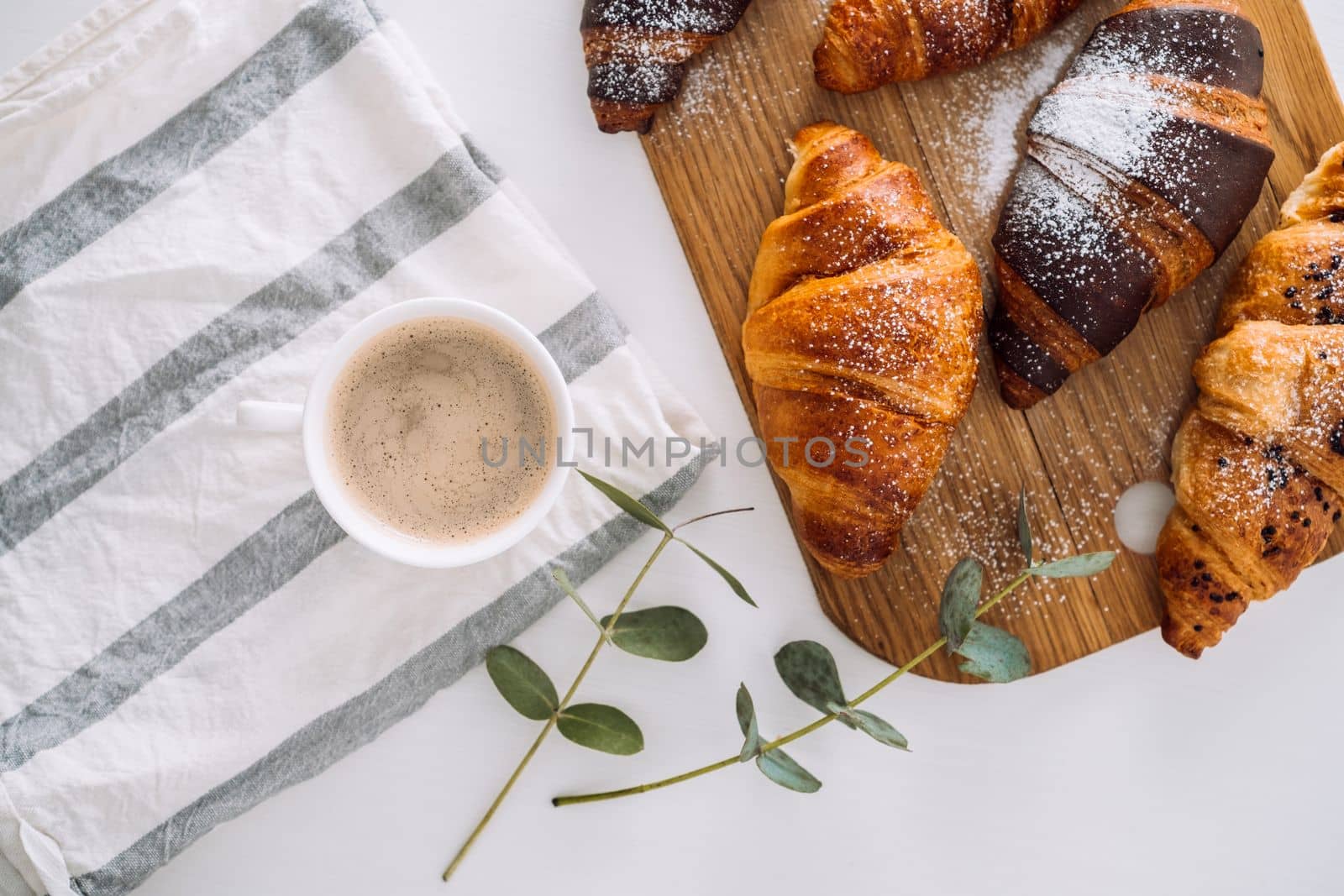 Cup of cappuccino with kitchen towel and eucalyptus branch, brown croissants on cutting board