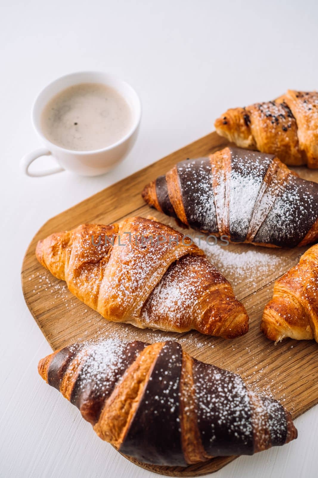 Bunch of appetizing brown and chocolate croissants with powdered sugar on a wooden board with cup of coffee on white table by Romvy