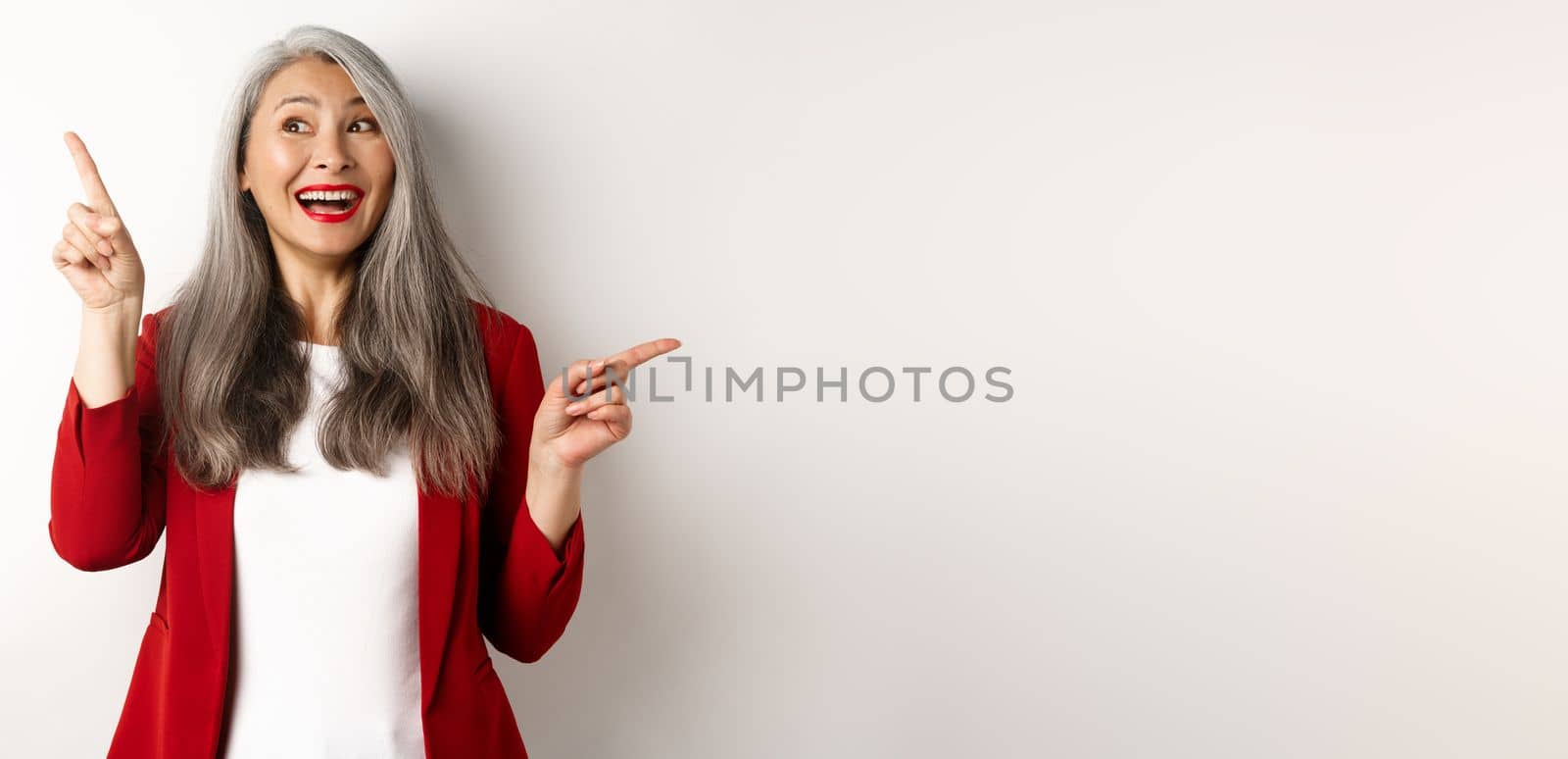 Cheerful asian lady smiling, pointing fingers sideways at two promo offers, standing happy in red blazer over white background.
