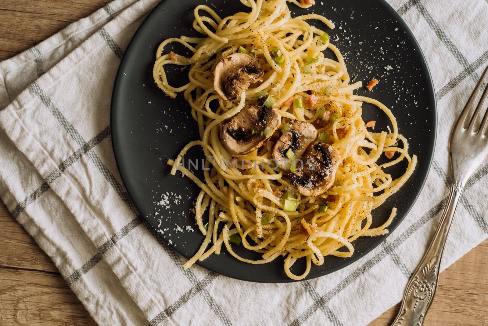Spaghetti pasta with champignon mushrooms sprinkled with cheese parmesan and green onions on a black plate with a towel and a fork on a wooden table by Romvy