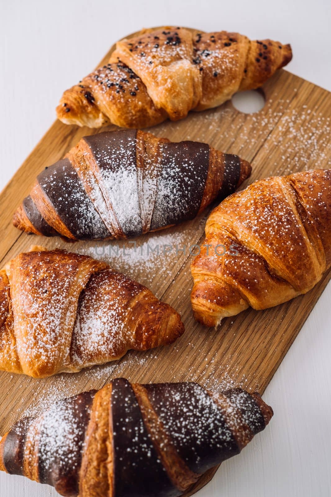 Close up bunch of appetizing brown and chocolate croissants with powdered sugar on a wooden board on white table by Romvy