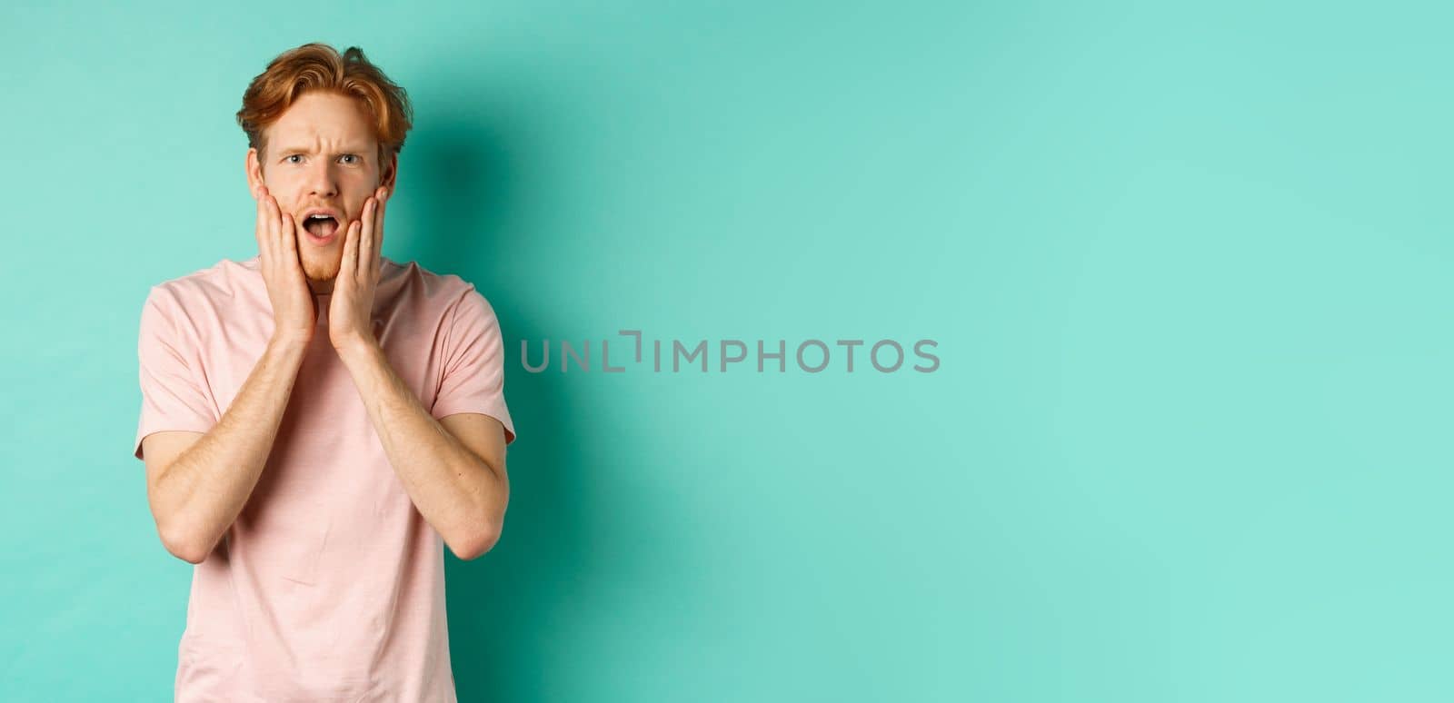 Shocked and concerned young man with red hair, staring at camera worried and touching face, standing in t-shirt against turquoise background.